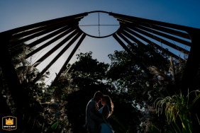 The couple stands close together in silhouette beneath a dramatic architectural structure against the afternoon light in Tulum, Mexico